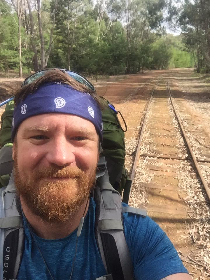 Jed with bead wearing a bandana with his big backpack on with a railway track behind leading off into distance