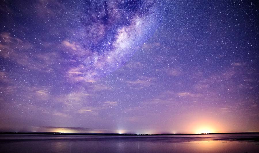 the milky way in a purple sky with lights from town over the horizon