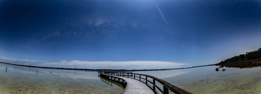 Blue sky with very faint milky way over Jetty over water. Clouds form a low line on the horizon 