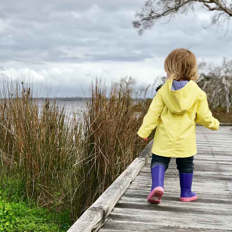 Maddie in a yellow rain jacket and purple gum boots walking along a boardwalk beside the estuary