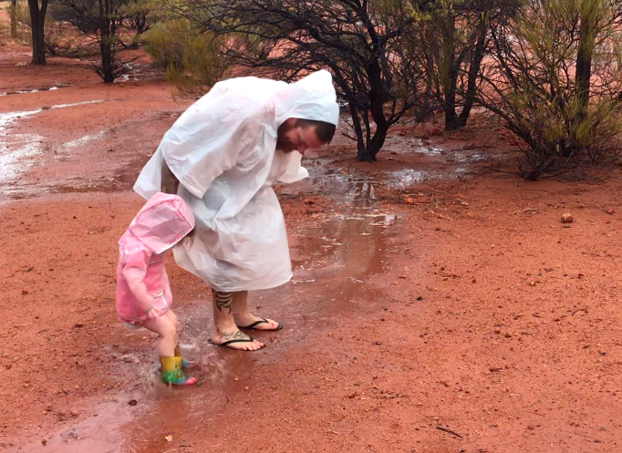 Jed wearing a plastic poncho and Maddie wearing her pink mac and rainbow gum boots jumping in a puddle